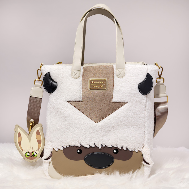 Plush tote bag that looks like Appa from Avatar: The Last Airbender, with embroidered and appliqué details with beige straps and a Momo charm sitting on a furry white blanket against a white background